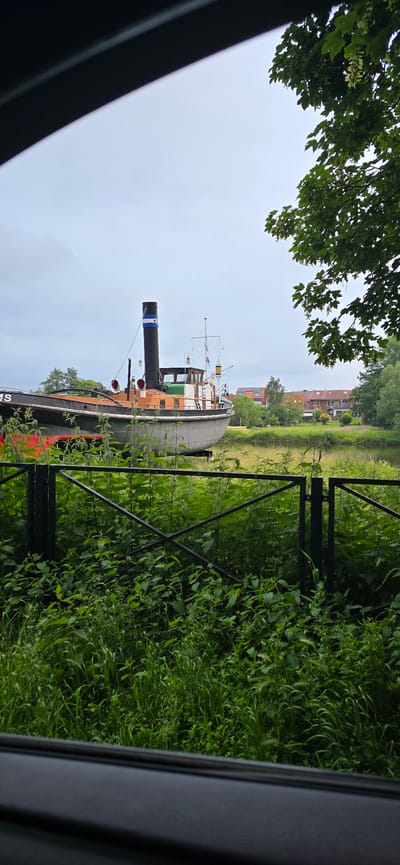 A tugboat, identified by lettering that appears to read "NS" on its smokestack, is beached and displayed on land. The tugboat is dark grey with an orange and green superstructure. A prominent black smokestack with blue and white banding rises from its deck. The boat sits on a grey, flatbed trailer.
The scene is viewed through a black frame, possibly from inside a vehicle, with the lower portion of the frame showing a dark, textured surface and the upper portion showing a sliver of a black frame and an overcast sky.
In front of the tugboat, a black metal fence runs horizontally across the frame. Behind the fence, lush green vegetation, including tall grass and nettles, grows abundantly. Beyond the vegetation and the tugboat, a calm body of water is visible, with grassy banks on the far side. Further in the distance, a cluster of residential buildings with red-tiled roofs can be seen nestled among trees. The overall impression is a serene, somewhat melancholic scene of a retired vessel resting on land in a tranquil, green environment, likely in Haren (Ems), Germany, given the context provided.