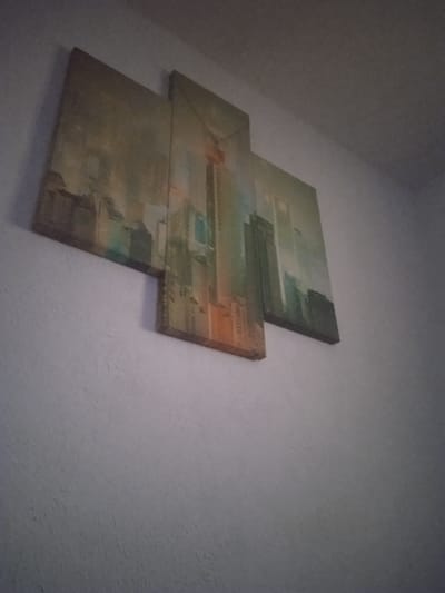 This image shows a three-panel canvas art piece depicting a city skyline, likely an abstract or impressionistic representation of skyscrapers. The art is mounted on a textured white wall. The colors in the artwork range from muted browns and grays to hints of orange and teal, suggesting a daytime or dusk scene. The perspective of the photograph is from below, looking up at the artwork. The environment appears to be indoors. There are no people or any other objects visible in the image besides the artwork and the wall. The location of Nihoreni, Moldova, is not identifiable from the visual information presented in the image.