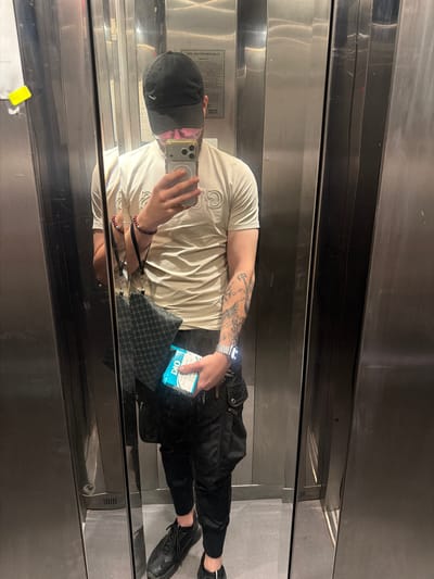 The image shows a person in an elevator, likely in the city of Kyustendil, Bulgaria, reflected in a mirrored wall. The person is wearing a black baseball cap, glasses, a light-colored t-shirt with a graphic design, black cargo pants, and black sneakers. They are holding a smartphone in their right hand, taking a selfie, and a package of what appears to be cigarettes in their left hand. A dark patterned pouch or bag is hanging from their left shoulder. The elevator appears to be modern, with stainless steel walls and a gray floor. The lighting is artificial, typical of an indoor environment. There are no other people or objects of significance visible in the frame.
