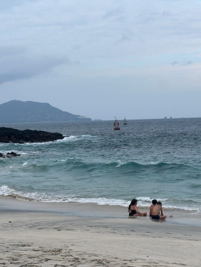 An outdoor, eye-level shot captures a serene beach scene with three individuals sitting on the wet sand near the water's edge. The person on the left, a woman wearing a black swimsuit, is seated facing towards the right with her back to the camera. Next to her, a boy in dark swim trunks is seated with his back to the camera, looking out at the sea. To the boy's right, another boy, also in dark swim trunks, is seated with his back to the camera. The ocean is a deep blue with gentle waves lapping at the shore, and two buoys are visible in the distance. A mountainous landmass is visible on the horizon. The sky is overcast with clouds. This image was taken in Manggis, Indonesia.
