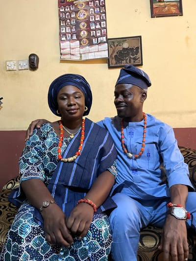 A photo shows a man and a woman indoors. The man is wearing a light blue shirt and a matching hat with blue stripes, and an orange beaded necklace. He has a wristwatch with a red band on his left wrist. The woman is wearing a dark blue top with blue stripes and a matching head wrap. Her top is patterned with white dots and blue embellishments. She also wears an orange beaded necklace and a bracelet on her left wrist. The two individuals are sitting close together. The woman is looking at the camera and smiling. The background is a yellow wall with two framed pictures and a calendar hanging on it. The location is Rafin Bauna, Nigeria, although this information cannot be visually confirmed from the image.