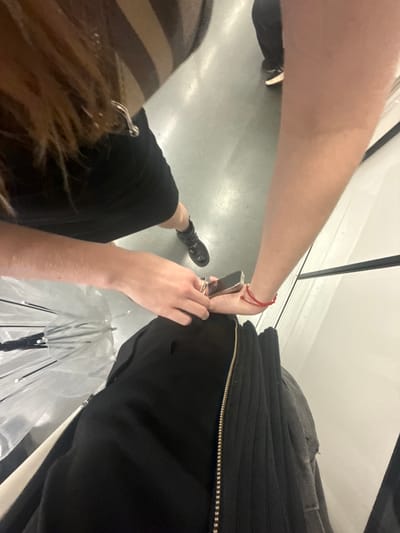 The media file shows a close-up, high-angle shot of two people in what appears to be an indoor, possibly public, setting. The primary subject is a person's arm and hand, which is reaching towards a zipper on black clothing. The arm has a red string bracelet on the wrist. The hand is holding a small, metallic object, possibly a lighter or a small tool, near the zipper.
The lower part of the frame reveals another person's legs and hair. One leg is bent, wearing black boots and black clothing. The other person's hair is visible, with a striped garment partially showing. A clear umbrella with black accents is positioned near the bottom right of the frame, suggesting it might be a personal item.
The ground appears to be a smooth, light-colored surface, possibly concrete or tile, with some subtle reflections. The background shows a white wall and a dark vertical element, suggesting an indoor environment. The lighting is bright, indicating daytime or well-lit indoor conditions. There is no visible text in the image. The overall mood is unclear due to the close-up nature of the shot and the focus on the action of interacting with the zipper. The setting is likely urban, and the activity appears to be casual or personal. The context of Changning District, China, suggests this is a scene within that geographical area.
