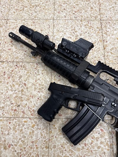 A black rifle with a scope and a red dot sight is laying on a tiled floor. A black handgun is positioned beneath the rifle. The tile floor is composed of small, irregular, cream-colored stones. The weapons appear to be in good condition. The location, based on coordinates (latitude: 31.833, longitude: 34.82), is in Mazkeret Batya, Israel.