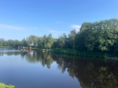 The image depicts a serene waterscape with a clear blue sky and reflections of trees on the water's surface. In the distance, a small yellow and red structure, possibly a small dock or bridge support, is visible on the water. The lush green trees line the bank of the body of water, creating a natural barrier. The overall scene suggests a tranquil natural environment, likely a park or a waterway within a town. This scene is likely located along the **Balsjer Fleet** in Edewecht, Germany, known for its picturesque waterways and green spaces.