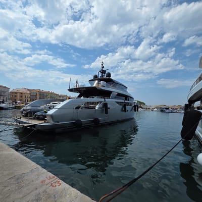 This media file captures a luxurious yacht docked in a marina, likely in Saint-Tropez, France, given the contextual information. The primary subject is a sleek, dark-grey yacht with modern architecture, featuring large windows and multiple levels. The yacht is moored alongside a concrete dock, with thick ropes connecting it to the shore. Other vessels are visible in the background, suggesting a busy harbor.

The scene is set during the daytime, under a bright blue sky dotted with fluffy white clouds, indicating fair weather. The water in the marina is calm, reflecting the sky and surrounding boats. Buildings with characteristic Mediterranean architecture, painted in warm hues of yellow and orange, line the waterfront in the distance, contributing to the picturesque coastal setting.

Several black fenders are strategically placed along the yacht's hull to prevent damage. The name "NEW YORK" is clearly visible on the stern of the yacht, along with a smaller text that appears to be "SACS". A small inflatable boat is stowed on the deck of the main yacht. A person or two can be seen on the deck of another yacht in the background.

On the concrete dock in the foreground, some red spray-painted markings are visible, possibly indicating dock numbers or mooring information. The overall impression is one of opulence and a relaxed maritime atmosphere.