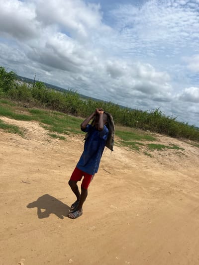 A young person, appearing to be a young male, is depicted walking along a dirt path in an outdoor, rural or semi-rural setting. They are wearing a blue short-sleeved shirt and red shorts, with black sandals on their feet. A large, possibly woven, bundle or bag is balanced on their head, and their hands are raised, appearing to stabilize the load.
To the person's left, a dense expanse of tall green vegetation, resembling crops or wild grass, borders the unpaved, light brown dirt path. The sky above is partly cloudy, featuring a mix of lighter, more scattered clouds and larger, denser formations. The sun, positioned high in the sky, casts a distinct, relatively short shadow of the person onto the dirt path, suggesting it is midday or early afternoon. The overall scene conveys a sense of daily activity in a natural environment. No visible text is present. This scene takes place in Idah, Nigeria.