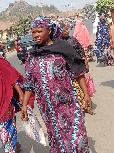 Several women wearing brightly patterned dresses and head coverings are lying on a paved surface, possibly a road. One woman holds a sack labeled "Dangote Cement". A black vehicle is partially visible in the background along with buildings and a hill in the distance under a clear sky. The scene appears to be outdoors in a busy area.