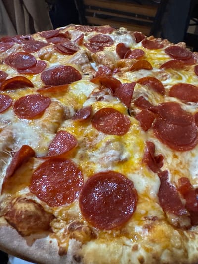 A close-up shot of a pepperoni pizza. The pizza is heavily topped with pepperoni slices and melted cheese. The crust appears to be slightly browned. The scene suggests the pizza is served indoors. No specific location details are discernible from the image, therefore the city location cannot be confirmed.