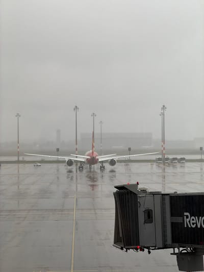The image depicts a wet airport tarmac in Schönefeld, Germany, under foggy conditions. Several passenger airplanes are parked in a row. An airport jet bridge labeled "Revo" is connected to one of the aircraft. Lighting poles, painted with white and red bands, stand alongside the planes. The ground is wet, reflecting the light and the shapes of the objects. Visibility is low due to the fog. There are no people visible in the frame.