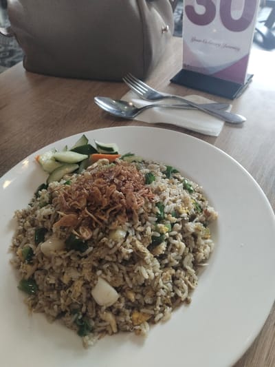 The image shows a plate of fried rice with a garnish of fried onions and cucumber slices. The fried rice is a mixture of white rice, green onions, and other unidentified ingredients. The dish is sitting on a wooden table in a restaurant, likely in Bekasi Regency, Indonesia, based on the coordinates provided. There are two spoons and a napkin to the right of the plate, and a table number holder to the right of the spoons. The plate is white, and the dish is a simple, everyday meal. There are no people in the image.