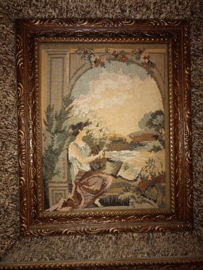 This is a framed tapestry depicting a pastoral scene. A woman in a light-colored dress and dark hat is seated on a bench, holding a basket and tending to plants. She is positioned under an arched structure with floral embellishments. Behind her, a body of water is visible, reflecting the sky. The tapestry is housed in an ornate, dark brown and gold frame. The tapestry and frame are set against a textured, patterned brown background, likely a wall or fabric covering. The image does not contain any identifiable landmarks or elements that would allow for localization in the city of Motmos, Russia.