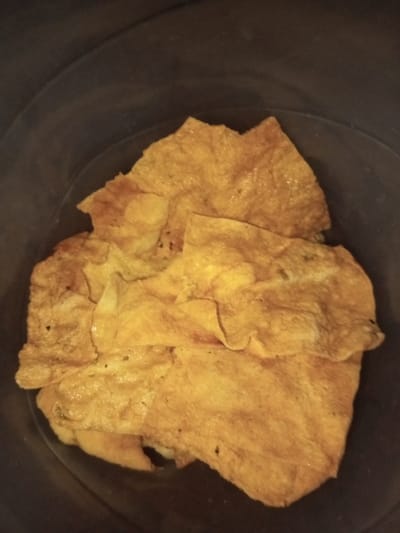The image depicts a pile of thin, golden-yellow omelets inside a dark container. The omelets appear to be slightly greasy with some dark specks, potentially pepper. They are folded and layered irregularly, suggesting they were casually placed inside. The container's dark interior provides a contrasting background to the omelets.