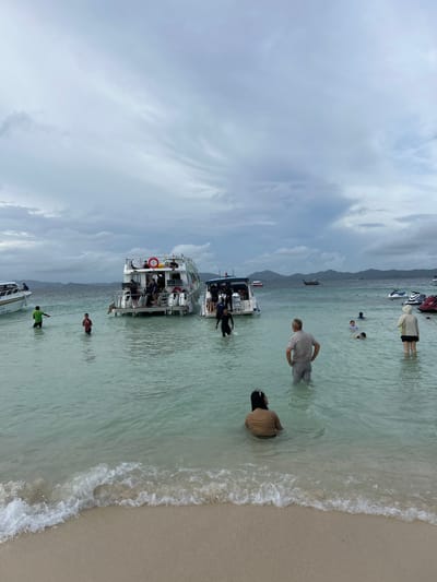 The image captures a beach scene with several boats docked in shallow, clear turquoise water under an overcast sky. In the foreground, a gentle wave breaks on a sandy shore. Several people are visible in the water, wading or swimming. Two white boats with multiple decks are anchored closer to the shore, with some individuals standing on their decks and others in the water around them. Further out, more boats, including a jet ski, are present in the distance. The horizon shows a line of hills or mountains. The overall impression is a popular tourist destination with recreational activities taking place. This scene is consistent with the coastal areas of Tambon Phru Nai, Thailand, known for its beaches and island hopping tours.
