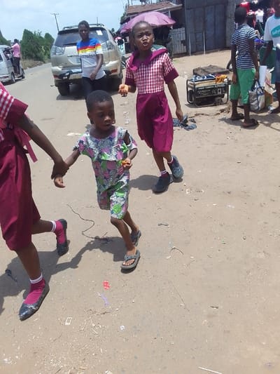 The image displays an outdoor street scene in Nsugbe, Nigeria, under bright daylight conditions. In the immediate foreground, three children are positioned on an unpaved, light-brown dirt road containing sparse debris. On the far left, a child, partially visible and wearing a maroon dress with a red-and-white plaid upper section, maroon socks, and black flat shoes, extends their left hand to hold the left hand of a second child. This second child, centrally located, wears a pink, green, and white patterned romper, grey flip-flops, and holds a small, orange-colored spherical object in their right hand. To the right of the central child, a third child, dressed in a maroon and red-and-white plaid school uniform with a white collar, dark athletic shoes, and white socks, holds two similar small, orange-colored spherical objects in their right hand while facing forward with their mouth slightly open.

In the mid-ground, a dark-colored portable generator is situated on the right side of the road. Behind the children, on the left, a light-colored SUV is parked, with an adult female wearing a white t-shirt featuring horizontal rainbow stripes and dark trousers standing nearby. Further into the background, an adult male in a striped t-shirt and light green shorts is visible on the right, standing near the generator. Additional individuals are partially visible throughout the background, including a person near a white van on the far left and another under a purple umbrella near simple commercial-type structures on the right. Utility poles and green foliage are discernible against the sky in the distant background.