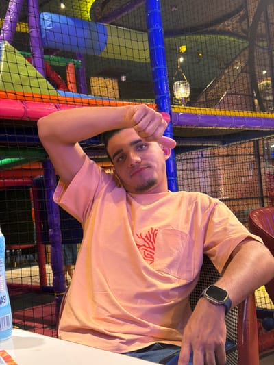 In this media file, a young man is captured in a playful, upside-down selfie. He's wearing a peach-colored t-shirt with a red coral-like design on the chest pocket and a black smartwatch on his left wrist. The background reveals a vibrant indoor playground, characterized by colorful padded poles and netting. Purple, blue, and yellow structures are visible, suggesting a soft play area for children. The lighting appears to be artificial, with some warm, possibly ambient light sources casting a glow on the scene.
The man is making a thumbs-up gesture, with a slight smile on his face, conveying a sense of fun and positivity. His eyes are open and looking towards the camera, and he has a light beard. The image seems to be taken at an indoor establishment, possibly during daylight hours given the artificial lighting, but without clear external cues. The overall mood is lighthearted and energetic, typical of a recreational activity. No visible text is present in the image itself.