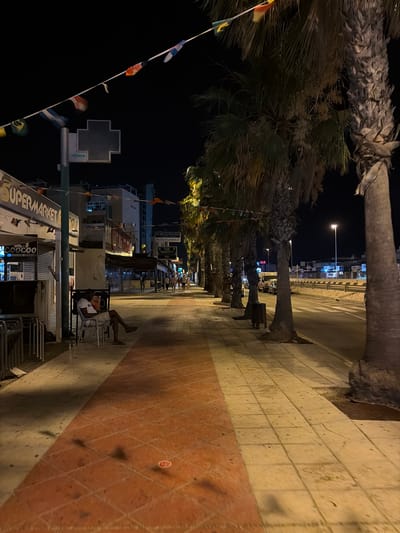 The image depicts a street scene at night, likely in a tourist area given the palm trees, streetlights, and numerous shops. A man sits on a white chair outside a closed "Supermarket" store, looking at his phone. He is wearing a white t-shirt and light-colored shorts. The pavement is a mix of light-colored square tiles and reddish-brown rectangular tiles. Small flags are strung across the street overhead. In the background, several buildings line the street, some with illuminated signs, and other pedestrians are visible walking further down the sidewalk. A road with a crosswalk runs parallel to the sidewalk, with streetlights illuminating the area. The overall impression is of a quiet evening in a commercial district.