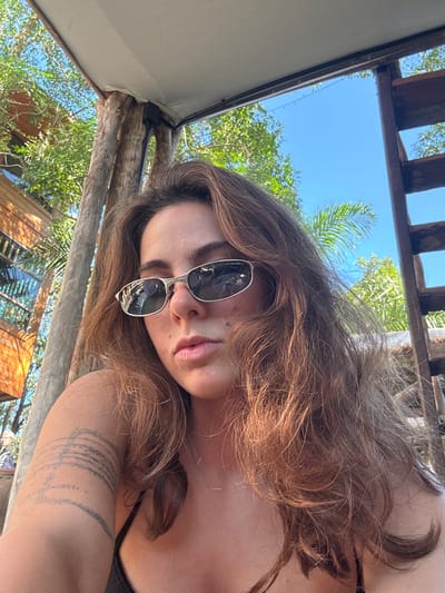 A woman with long brown hair and sunglasses poses outdoors. She is under a wooden structure with a partial white awning. Through the structure's openings, green trees and a bright blue sky are visible. A tattoo adorns her upper arm. Part of a building is visible in the lower left corner.