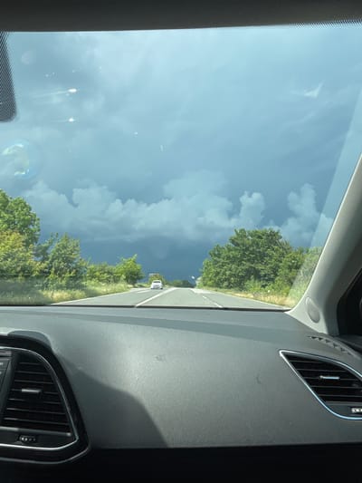 A forward-facing view from inside a moving vehicle on a road located in Bliznatsi, Bulgaria. The scene depicts a paved road extending into the distance, with a distinct central reservation and white lane markings. Dense green trees and shrubs line both sides of the road. The sky transitions from lighter, scattered clouds in the mid-ground to dark grey, heavy cloud formations in the far distance, suggestive of impending weather. A white van or car is visible travelling in the same direction, far ahead on the road. The lower portion of the image displays the vehicle's interior, specifically the dark grey, textured dashboard, which includes a portion of an air vent with metallic trim on the left and a similar vent partially visible on the right. The interior trim of the right A-pillar, featuring a circular speaker grille, is also visible. The windshield exhibits several reflections, notably a prominent circular, iridescent reflection in the lower left quadrant and multiple smaller, elongated light reflections scattered across the upper section of the glass. No people are present in the image.