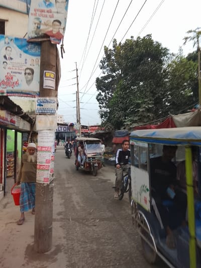 An outdoor scene depicts a street in a commercial area of Tarabo, Bangladesh. In the immediate foreground, a weathered utility pole, heavily adorned with Bengali-language election posters and various advertisements, dominates the left side. An elderly man with a long white beard, wearing a light brown shirt, blue checkered lungi, and sandals, walks past the pole toward the left, carrying a red plastic bucket in his left hand. To his left, a shop with a corrugated metal awning displays items internally.
The unpaved or worn asphalt road extends from the midground into the distance, populated by various vehicles. A blue and white three-wheeled auto-rickshaw is partially visible in the right foreground, moving away from the viewer, with an occupant visible inside. Ahead of it, a young male wearing a black long-sleeved Adidas shirt and light pants rides a bicycle, also moving away. Further down the road, a reddish-brown three-wheeled motorized vehicle with multiple occupants is present, followed by a person on a motorcycle and other blurred figures.
Along the right side of the road, green trees rise above a series of makeshift stalls or shops with red and beige/grey tarpaulin roofs. Utility poles and a network of overhead power lines crisscross the sky, which appears overcast or hazy. Buildings of varying sizes, including one marked "ALTON," line the street, indicating a bustling urban environment.