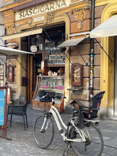 The image depicts an outdoor scene in Ljubljana, Slovenia, featuring a building identified by the sign "SLASCICARNA." The facade of the building shows ornate carvings and detailing above the entrance, with several framed pictures hanging on the wall, as well as handwritten signs advertising various food and drink options. A white bicycle with a child seat attached to the back is parked to the side of the building, partially obscuring the entrance. A black plastic chair is also visible on the cobblestone street. The photograph is taken at an angle, causing a slightly distorted perspective of the scene.