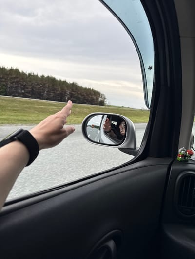 The image, taken from inside a car, shows an arm extending out of the window. The wrist has a black watch band. The hand appears to be waving. The side mirror reflects a person wearing glasses, also waving. Outside, the landscape consists of a road flanked by a field or grassy area and a treeline. The sky is overcast. In the car's interior, a small decorative object with multiple figures is attached to the car's dashboard. The city is not identifiable in this image.
