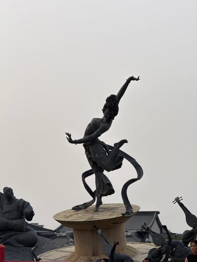 This image shows a statue in Lintong District, China. A bronze statue of a female figure, in mid-air, is supported by a circular base. The figure is posed in a dance-like posture with one arm raised, wrapped in flowing fabric. Around the base of the central figure are other smaller statues playing stringed instruments, with Chinese architecture visible in the background. The sky overhead is overcast.