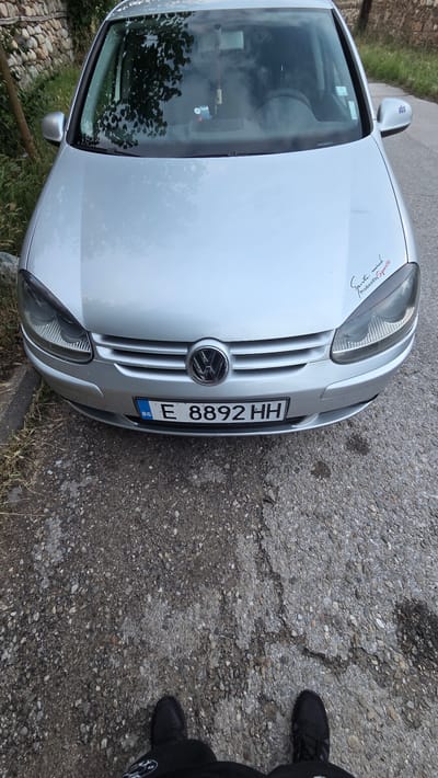 The image shows a silver Volkswagen Golf parked on a paved road. The license plate reads "E 8892 HH" with "BG" indicating Bulgaria. A sticker on the hood says "Sports mind. Produced by Sports". The car is facing the camera, and the lower part of the image shows a person's feet wearing black sneakers. The background includes a stone wall and some greenery. The scene is likely in the city of Bania, Bulgaria, given the license plate.