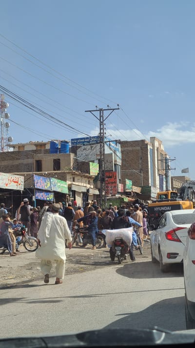 This image captures a vibrant and bustling street scene in Kuchlak, Pakistan, under a clear, bright blue sky with scattered white clouds, indicating daytime. The perspective is from inside a vehicle, looking out onto a busy road.

Numerous people, predominantly men dressed in traditional South Asian attire like shalwar kameez, fill the street. Many are gathered or waiting, while others are seen riding or standing next to motorcycles. One man in a light-colored shalwar kameez walks away from the camera on the right side of the road. A white sack, likely containing flour or grains, is strapped to the back of a motorcycle in the mid-ground.

The street is lined with multi-story buildings that appear to be commercial establishments, featuring a mix of exposed brickwork and modern glass facades. These buildings are adorned with many colorful signboards, primarily displaying text in Urdu, indicating various businesses or advertisements. A prominent utility pole with a dense network of overhead wires stands on the left side of the street, extending across the frame.

Vehicle traffic includes several motorcycles and at least two cars. A white car, possibly a sedan, is parked or moving slowly on the right, and behind it, part of a large yellow heavy vehicle, likely a construction machine like an excavator or loader, is visible, with the brand "HYUNDAI" clearly written on its side. The road surface is asphalt, and shadows are visible on the ground, suggesting bright sunlight. The overall atmosphere is one of a busy, crowded commercial area.