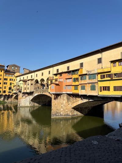 The Ponte Vecchio bridge in Florence, Italy, is captured from a low angle, showcasing its iconic medieval structure spanning the Arno River. The bridge is lined with shops and buildings painted in vibrant yellow and orange hues, with arched openings revealing the stonework beneath. The clear blue sky above is dotted with a few birds in flight. The river below reflects the colorful buildings and the stone arches, creating a shimmering effect on the dark green water. The foreground features a dark, textured surface, possibly a stone embankment or pathway, leading the eye towards the bridge. The scene is bathed in bright sunlight, suggesting it is daytime.