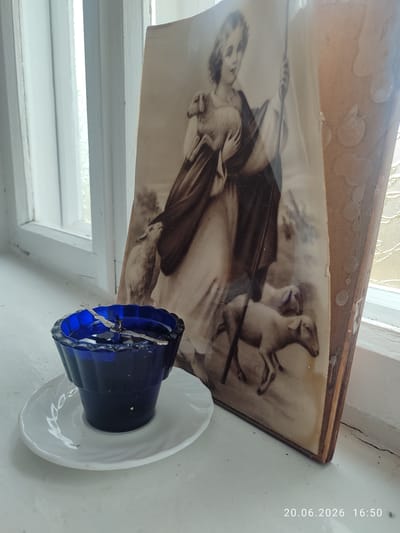 A close-up, eye-level shot captures a devotional scene on a white windowsill in Tomai, Moldova. In the foreground, a small, ornate blue glass candle holder, filled with what appears to be lamp oil or water, sits on a white saucer. A metallic wick holder with a wick is submerged in the liquid. To the right and slightly behind the candle holder stands a framed sepia-toned print depicting a young boy, possibly Jesus, holding a lamb. The boy is dressed in flowing robes and carries a staff. Several other sheep are visible in the background of the print. The print is leaning against the windowsill, its wooden backing aged and showing signs of wear. The overall environment is dimly lit, with light filtering through a nearby window, creating highlights on the objects. A date and time stamp, "20.06.2026 16:50", is present in the lower right corner of the image.