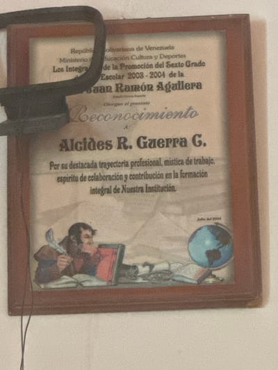 A framed certificate is displayed on a white wall, with a dark border and wooden frame. The certificate is cream-colored and features text in Spanish, along with an illustration. At the top, the text reads "República Bolivariana de Venezuela Ministerio de Educación Cultura y Deportes". Below this, it states "Los Integrales de la Promoción del Sexto Grado Escolar 2003-2004 de la". Then, a large, bold name is visible: "Juan Ramón Aguilera". Further down, the text "Otorgan el presente Reconocimiento" is present. The main name on the certificate is "Alcides R. Guerra G.", written in a large, bold font. Below this, more text describes "su destacada trayectoria profesional, mística de trabajo, espíritu de colaboración y contribución en la formación integral de Nuestra Institución." In the lower section of the certificate, an illustration depicts a man with a quill pen in his right hand, seated at a desk with an open book and a red object that appears to be a book or a box. To the right of the illustration is a small globe and some rolled-up papers. The bottom right corner of the certificate has the text "Julio del 2004". The overall lighting suggests an indoor setting, possibly a room within El Maco, Venezuela, given the information on the certificate.