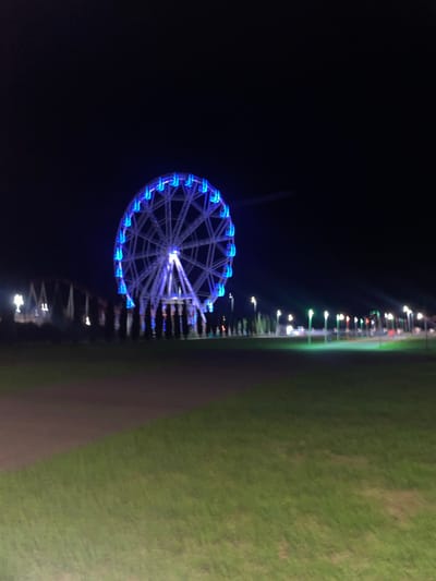 The image depicts a Ferris wheel illuminated with blue lights at night in Gagra, Abkhazia. The Ferris wheel is the central and largest object in the frame, dominating the left side of the image. It is a large, circular structure with spokes and carriages, all outlined by bright blue LEDs. To the left of the Ferris wheel, out of focus, are some buildings or structures with lights, possibly part of an amusement park or resort. In the foreground, there is a grassy area with a path or walkway. Beyond the Ferris wheel, to the right, a line of streetlights or park lights with various colored glows (green, white, red) stretches into the distance, receding into the dark sky. The overall environment suggests a recreational area, likely a park or an entertainment complex, at night. There are no people visible in the image. The scene is characterized by the contrast between the dark night sky and the bright, artificial lights of the Ferris wheel and other illuminated elements.
