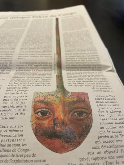 The image displays a close-up, angled view of an open newspaper laid flat on a dark surface in Cucuron, France. Dominating the center of the newspaper is a stylized illustration, cut into the shape of a shovel. The shovel's handle, extending upwards, features abstract patterns resembling colorful topographical maps or veins. The spade-like base of the shovel depicts a human face, rendered with a rich palette of oranges, browns, reds, and purples, incorporating map-like textures into the facial contours. Two distinct eyes are visible, along with the nose and mouth. The surrounding newspaper text is in French, with legible sections discussing the Democratic Republic of Congo (RDC), the "guerre froide" (Cold War), the assassination of Patrice Lumumba on January 17, 1961, and the involvement of Belgium and the United States. The overall lighting suggests an indoor setting.