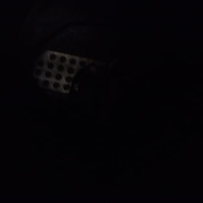 A dimly lit image reveals a partially visible, rectangular object with a grid-like pattern of circular perforations. The object's edges are indistinct, merging into the dark background. A shadowed, rounded form rests partially atop the gridded surface, further obscuring the details of the underlying structure. The overall scene is characterized by low visibility and limited detail.