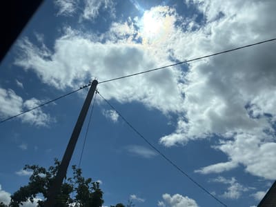 The image depicts a bright, partly cloudy sky over Капыльскі сельскі Савет, Belarus. A solitary utility pole, made of concrete and angled towards the left, stands prominently on the left side of the frame. Several wires are suspended from the pole, extending across the sky. The lower portion of the frame shows the upper branches of a tree with dark green foliage, partially obscuring the base of the utility pole. In the upper right corner, the edge of a brick building is visible. The sun is positioned in the upper central portion of the image, partially obscured by clouds, creating a lens flare and a subtle rainbow-like halo. The sky is a vibrant blue, with fluffy white clouds scattered throughout. The scene appears to be outdoors during daylight hours.