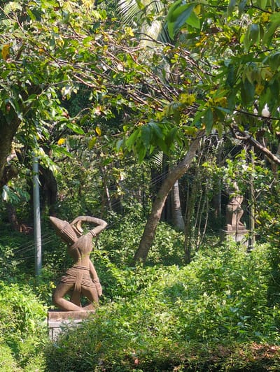 The image captures a scene in Tuy Hòa, Vietnam, focusing on a weathered stone statue of a figure in a natural setting. The statue, which has traditional clothing and ornamentation, stands among lush greenery. Other statues can be seen in the distance, creating an impression of a statue garden or area where traditional figures are displayed. The setting is outdoors, with trees and plants dominating the environment, and utility lines visible in the midst of vegetation.