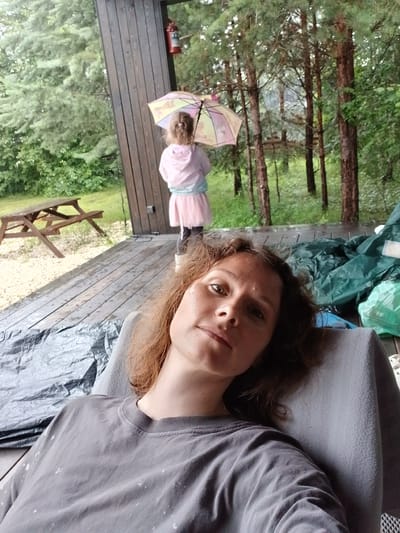 A woman is relaxing on a porch overlooking a wooded area. She is wearing a gray t-shirt. In the background, a young child is standing under an umbrella, facing away from the camera. The child is wearing a pink hooded jacket and a pink skirt. A red fire extinguisher is mounted on the wooden structure of the porch. The porch is wet, indicating it has been raining. A picnic table is visible on the gravel ground to the left. The overall setting appears to be a rural or semi-rural location, potentially a dacha or a house with a large yard. The city of Verkhneye Dubrovo, Russia, is mentioned as the location.