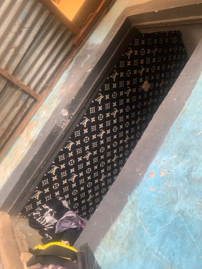 An overhead, tilted photograph captured in Bwari, Nigeria, depicts a closed, coffin-shaped object or large rectangular container positioned lengthwise within a narrow concrete doorway or alcove. The object is draped in black material featuring a repeating white "LV" monogram and quatrefoil pattern, secured by a single metallic latch or padlock positioned centrally along its visible length. Below the object, to the lower left, a disordered stack of various garments, including items in yellow and black, is visible. The immediate environment includes rough concrete walls, with surfaces painted in faded light blue and dark grey. Above and to the left, a corrugated metal roof structure supported by wooden beams, and a section of wire mesh are partially visible. The scene is illuminated by ambient daylight. No human subjects or discernible activity are depicted.
