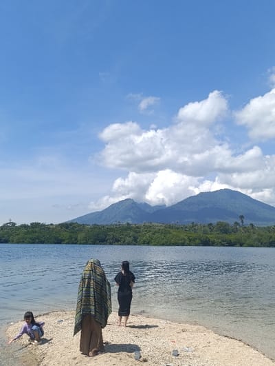 The image shows three people standing on a small sandy beach in front of a body of water, possibly a lake or estuary. The water is calm and the sky is clear blue with scattered white clouds. The people are facing away from the camera. The person on the left is wearing a brown headscarf and a brown long-sleeved dress, and the person in the middle is wearing a black dress. The person on the right is a young girl wearing a purple shirt and blue pants. The beach is covered in small white pebbles. In the background there is a lush green forest line, and behind that, a mountain range. The location is a coastal area in Kecamatan Banyuputih, Indonesia.