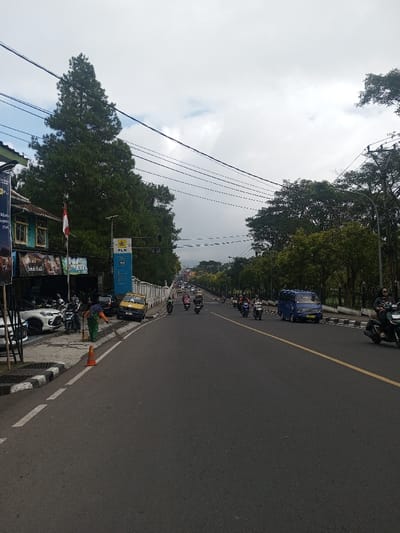 This image captures a daytime street scene, likely in an urban or suburban area with a somewhat hilly or mountainous backdrop under a cloudy sky. The perspective is from the side of a road, looking down a long, slightly curving street.
On the left side of the image, there's a sidewalk adjacent to a two-story building. The building has a green awning over what appears to be a shop or restaurant, with a red and white flag (possibly Indonesian) hanging from it. Below the awning, banners with text, some of which reads "Sari..." and "Sambal...," are visible. Several motorcycles and at least one white car are parked along the sidewalk in front of the building. A person wearing a green top and pants, possibly a street cleaner or worker, is sweeping the curb with a broom near an orange traffic cone.
The main subject of the image is the road itself, which has two lanes visible in this direction, separated by a solid yellow line. The road appears to be gently sloping downhill or curving. There's a fair amount of traffic, predominantly motorcycles, but also a blue minivan or small truck on the right lane, and a yellow car parked partially on the curb further down on the left. Many motorcyclists are visible, some individually, others in small groups, all heading in the same direction as the blue minivan.
On the left side of the road, beyond the parked vehicles and the sweeping person, a blue sign with "PLN" (an acronym often associated with electricity companies in Indonesia) and a lightning bolt logo is prominent. Tall evergreen trees line this side of the street further down.
On the right side of the road, there's a white concrete barrier or fence running along the curb, separating the road from what appears to be a green, treed area or park. More tall, leafy trees are visible lining this side of the street, extending into the midground. Utility poles and a web of overhead power lines or communication cables crisscross the upper part of the sky.
In the far background, the road continues, curving slightly, and distant hills or mountains can be seen under the overcast sky. The overall weather seems to be cloudy but not raining, suggesting a mild day. The scene depicts typical daily activity in what appears to be an Asian city or town.