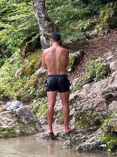 A person with dark hair, facing away from the camera, stands on a rocky surface next to a body of water, likely in Vusanje, Montenegro. The person is wearing black shorts. Their arms are crossed in front of their chest. To the left of the person, a grey athletic shoe is visible on a large rock. The background features a dense forest with trees and moss-covered rocks, indicating an outdoor, natural setting. The water appears calm and reflects the surrounding greenery.