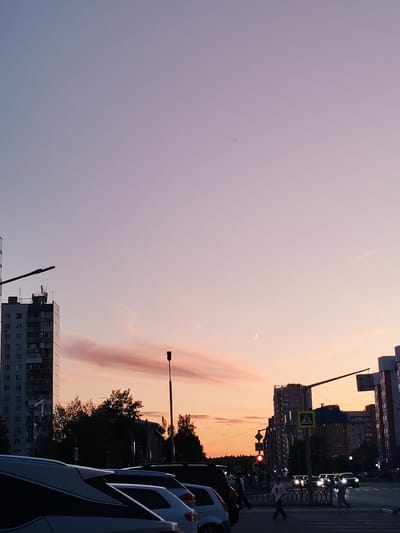 The image captures a street scene in Nizhnevartovsk, Russia, during twilight. A sky of soft pink and pale purple hues dominates the upper portion, with a crescent moon visible in the upper right quadrant. In the foreground, several cars are parked in a lot, with their rear ends visible.
The middle ground of the image features a street with traffic and pedestrians. Traffic lights are red, indicating a stop. Several buildings of varying heights line the street, including a tall apartment building on the left. Trees, silhouetted against the colorful sky, are interspersed between the buildings. Pedestrians are crossing the street at a marked crosswalk. The overall impression is of a typical urban environment at the end of the day.