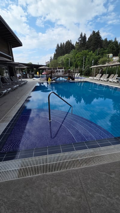 This is a medium-shot, eye-level, vertical photograph of a swimming pool area on a sunny day, likely in Bania, Bulgaria. The pool is large and filled with clear blue water, reflecting the sky and trees. The shallow end of the pool features blue tiled steps with a metal handrail. Surrounding the pool is a gray tiled deck with metal drainage grates. To the left, a building with a wooden facade and a dark roof is visible, with several lounge chairs and umbrellas set up outside. In the background, a wooden bridge crosses over a section of the pool, and lush green trees line a hill, suggesting a natural, outdoor setting. The sky is partially cloudy with scattered white clouds. No people are clearly visible in the image.