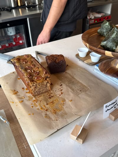 The image displays a food service counter in a establishment located in Bordeaux, France, likely a cafe or bakery. In the foreground, a brown paper liner covers a light-colored countertop. Resting on the paper is a partially sliced loaf cake, likely banana bread, topped with a caramel glaze and whole pecans. A slice has been removed from one end, leaving crumbs and drips of caramel on the paper. To its right, a small, dark chocolate cake, possibly a "fondant au chocolat," sits intact. A pair of metal tongs rests between the two cakes.

To the right, on a multi-tiered wooden stand, two onigiri (rice balls wrapped in nori) are visible on the top tier. Below them, two small, empty white ceramic ramekins are placed on a small wooden cutting board. Underneath the wooden stand, a stack of magazines or brochures is present. A small wooden block holder with a white card displaying "Fondant chocolat 4.10" is positioned near the right edge of the counter. Another similar wooden block holder with an empty white card stands further left.

In the background, a person wearing a dark vest or jacket and dark trousers is partially visible behind a stainless steel counter and equipment. Their right arm is visible. Behind them, a commercial refrigeration unit displays white and red cartons, and other kitchen equipment, including a red heating element, is visible on the back counter.