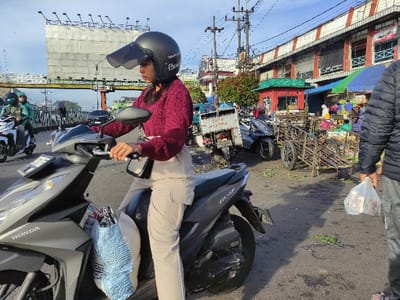 The image shows a woman wearing a red sweater and a helmet, riding a grey Honda motorbike on a paved road. She is holding a bag in her left hand and is looking to her left. The motorbike is parked on the side of the road, near a market with stalls and several carts. There is a billboard to the left side of the image. The image was captured in the city of Kecamatan Lawang, Indonesia, based on coordinates (latitude: -7.83, longitude: 112.697). There is a man walking in the background wearing a blue jacket and holding a plastic bag in his right hand. The sky is clear, and there is a building in the background.
