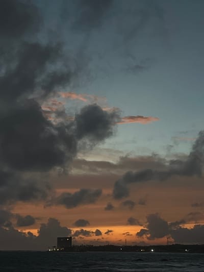 This media file is a photograph of a dramatic sunset over the ocean, likely taken from a coastal location in Galle, Sri Lanka. The composition is dominated by the sky, which displays a striking contrast between dark, brooding storm clouds and streaks of vibrant orange and yellow light, indicative of the sun setting. The clouds are textured and appear to be moving, creating a sense of dynamism.
The lower right portion of the image shows a sliver of the ocean, its surface ruffled with waves, suggesting a breeze or rougher seas. Along the horizon, a distant shoreline is visible, marked by a few scattered lights from buildings, suggesting an urban or semi-urban area. One prominent dark rectangular structure, possibly a building or a pier, is silhouetted against the illuminated sky. There are no people or discernible objects in the foreground or mid-ground, focusing the viewer's attention entirely on the natural spectacle of the sky.
The time of day is clearly sunset, characterized by the warm, atmospheric lighting and the fading daylight. The weather appears to be dynamic, with storm clouds gathering, yet the sunset's colors breaking through offer a sense of awe and perhaps a touch of melancholy. The overall mood conveyed is one of natural beauty, power, and a sense of impending change as day turns to night. No text is visible in the image.