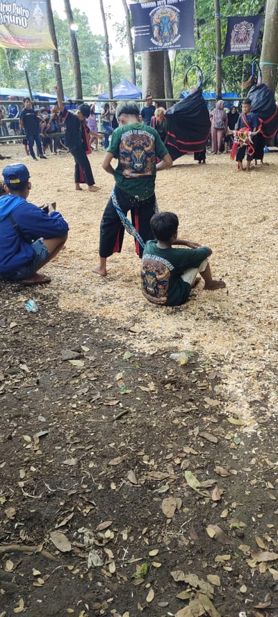 The image shows an outdoor scene in a park, likely in Kecamatan Lawang, Indonesia, with the given coordinates. There are a group of people in the scene, some wearing traditional Indonesian clothing. Two people in the foreground are wearing green shirts with a logo that says "ANNISA OF BANTENGANS". The people are standing on a dirt path with brown leaves scattered about. There is a black tent in the background, and a sign that says "BANDORO JAYA AUTO". The scene is likely during a public event or gathering. There are some items of litter on the ground including a plastic bottle and plastic bag. There are trees in the background and a metal ring in the upper middle of the picture.