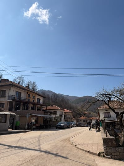 The image depicts a daytime scene on a street in Gradezhnitsa, Bulgaria, under a clear blue sky with a single white cloud. The street, a paved asphalt road, curves slightly to the right, receding into the background. Shadows from overhead utility lines and an unseen object are cast diagonally across the road surface from the left.

On the left side of the street, a multi-story building with a light-colored facade and dark roof tiles features balconies and an outdoor seating area with tables and chairs. Several individuals are present near this building, with at least two seated at a table. A green public telephone booth is visible adjacent to the building's entrance.

Along the right side of the street, a raised concrete sidewalk is bordered by a low, rough-hewn stone wall. A bare-branched tree is partially visible on the far right. Further down the street, a row of parked cars is visible, including a dark SUV. A group of approximately four to five individuals stands on the sidewalk to the right, engaged in an apparent interaction or observation.

Beyond the immediate structures, additional residential-style buildings with light facades and red-tiled roofs are situated. The background consists of rolling hills and mountains, largely covered with bare deciduous trees, indicating a season other than full summer, with some patches of evergreen foliage. Multiple black utility lines traverse the upper portion of the frame.