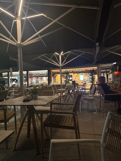The image shows an outdoor seating area of a restaurant complex at night in Keramoti, Greece. Multiple umbrella-like structures with integrated LED lighting provide overhead cover. In the background, several establishments are visible, including "MENOY" and "PIZZA PARADOX." The seating area is furnished with numerous tables and chairs made of wicker and wood. A few patrons are seated at tables or walking through the area. One man is visible sitting at a table in the foreground, facing away from the camera. Another person's arm is visible in the right foreground. The ground is paved with square tiles.