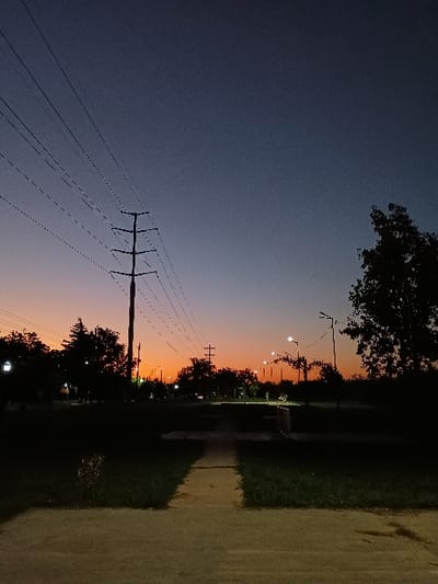 The image depicts a serene twilight scene in General Pacheco, Argentina, characterized by a striking gradient across the sky. The upper left portion of the sky is a deep, dark blue, gradually transitioning to a vibrant orange and yellow along the horizon line towards the right.
In the foreground, a prominent utility pole stands on the left, featuring multiple horizontal crossbars from which numerous thin power lines stretch across the mid-ground, forming intricate patterns against the colorful sky. A continuous silhouette of trees and dense foliage lines the horizon, suggesting a park or a tree-lined street. Several streetlights are visible within this area, their glowing bulbs emitting warm light that pierces the fading natural light.
To the right, a light-colored paved or dirt path runs parallel to the line of trees and streetlights, appearing to be a sidewalk or a trail. Shadows from the trees and possibly the utility pole are cast onto this path.
The scene is devoid of people or discernible activity, conveying a sense of quiet and stillness at the end of the day. The sky appears clear, with no clouds. No visible text is present in the media file.